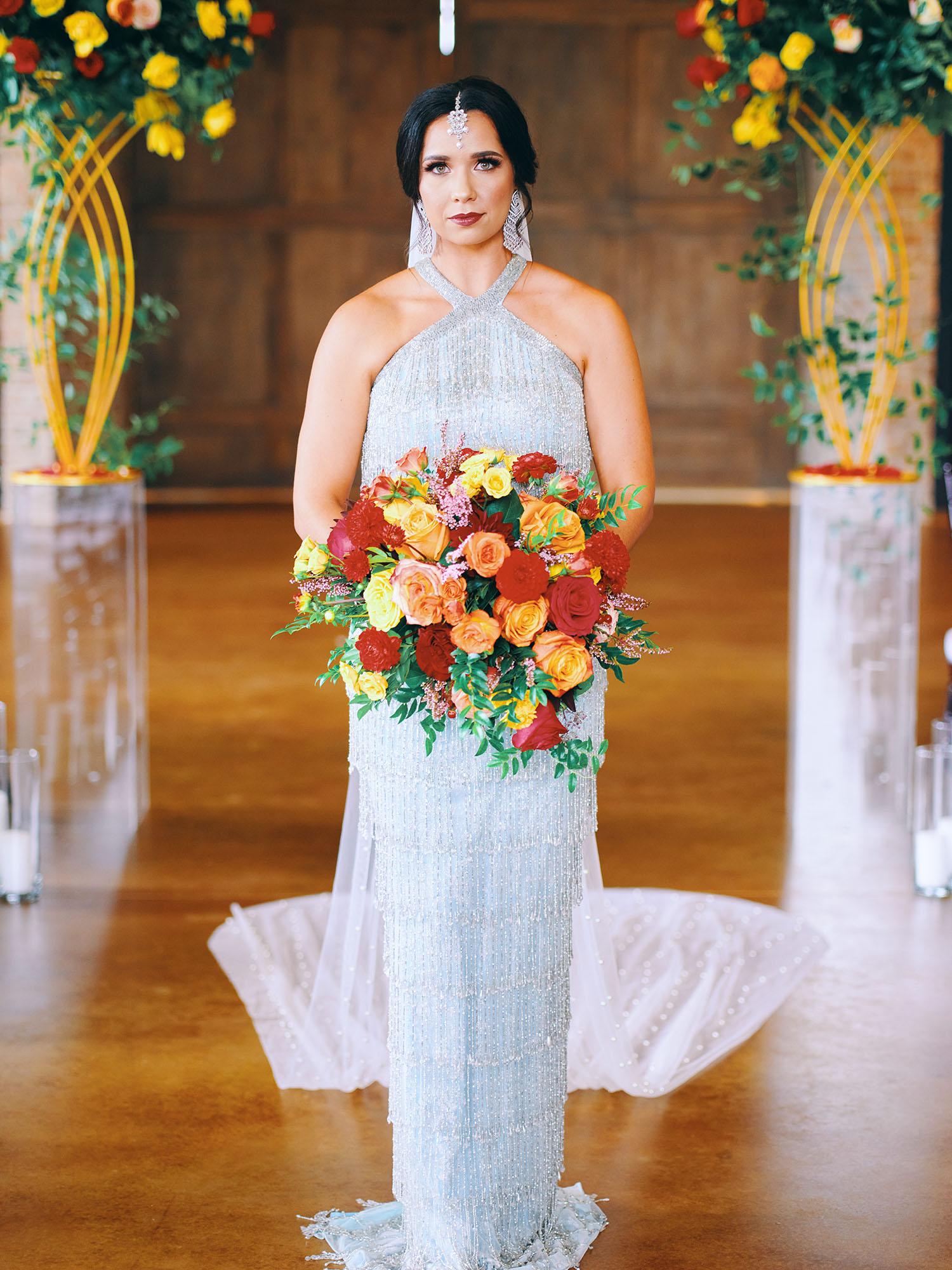 luxurious indoor wedding ceremony decor design, with a large red, yellow, and orange floral designs and a colorful bridal bouquet.