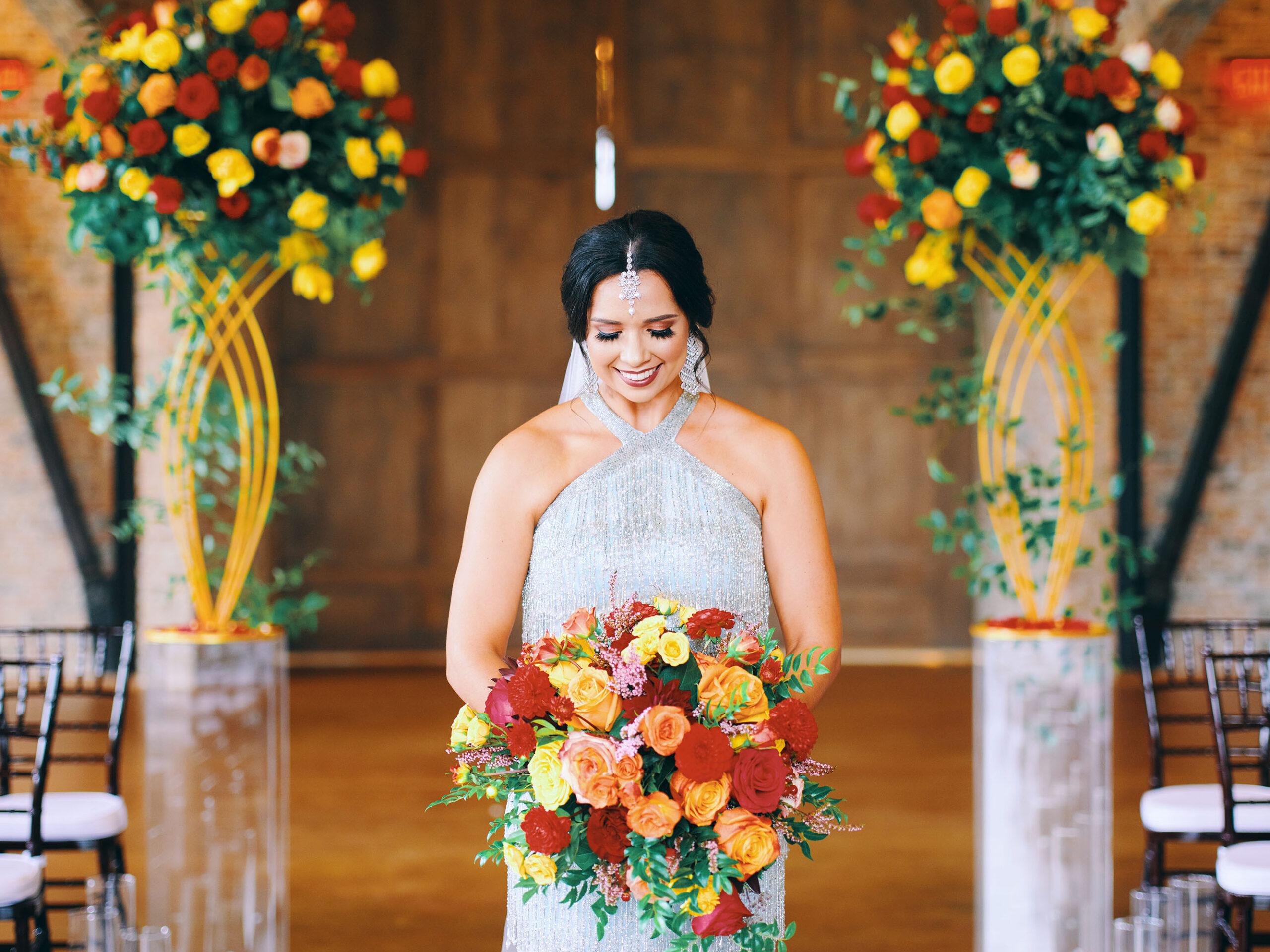 luxurious indoor wedding ceremony decor design, with a large red, yellow, and orange floral designs and a colorful bridal bouquet.