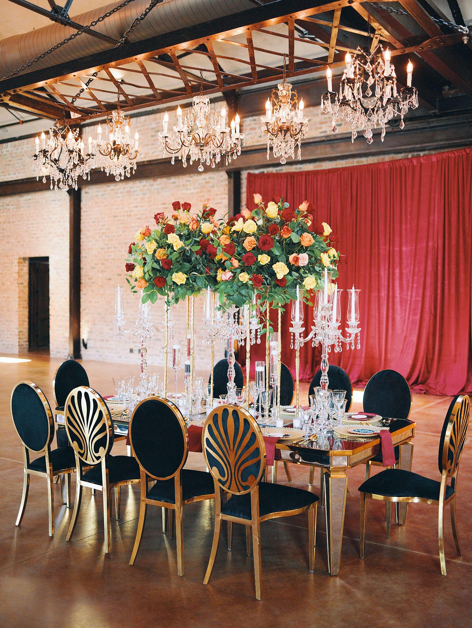 luxurious indoor wedding reception decor design, with red, yellow and orange floral designs, gold and black chairs, rectangular table, and a stunning bride and bridal bouquet.