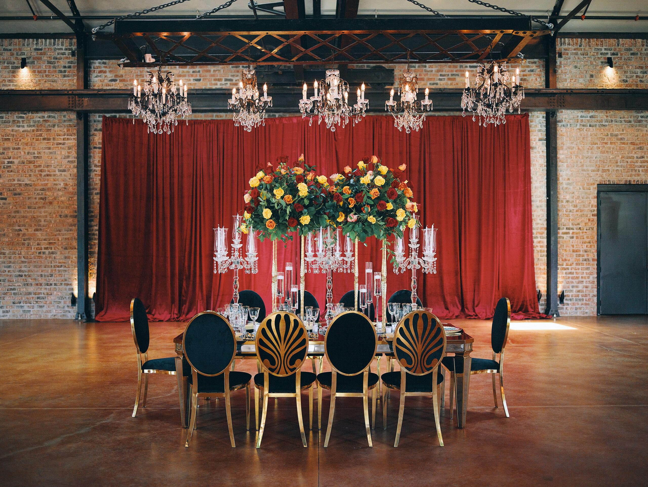 luxurious indoor wedding reception decor design, with red, yellow and orange floral designs, gold and black chairs, rectangular table, and a stunning bride and bridal bouquet.