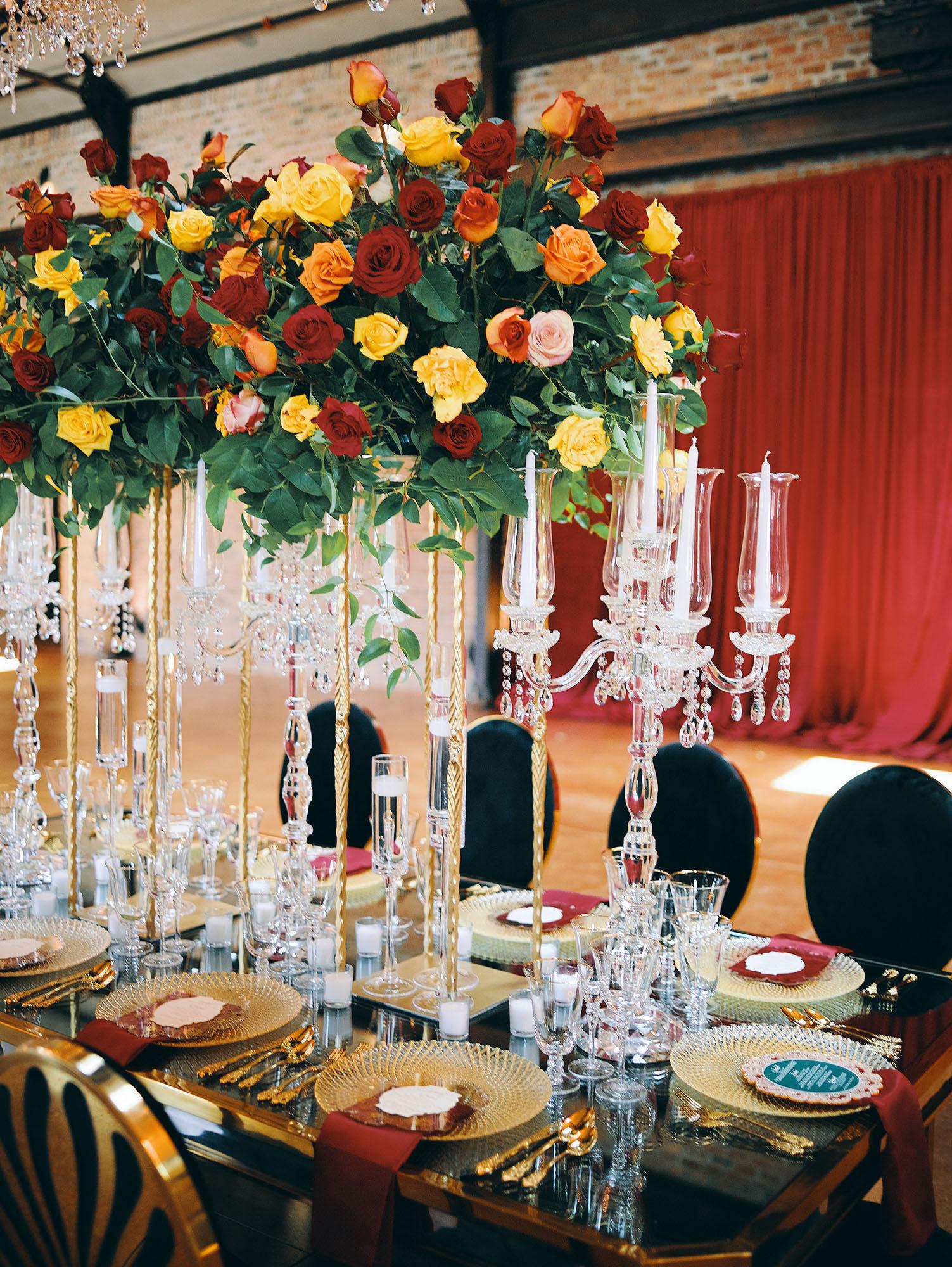 luxurious indoor wedding reception decor design, with red, yellow and orange floral designs, gold and black chairs, rectangular table, and a stunning bride and bridal bouquet.