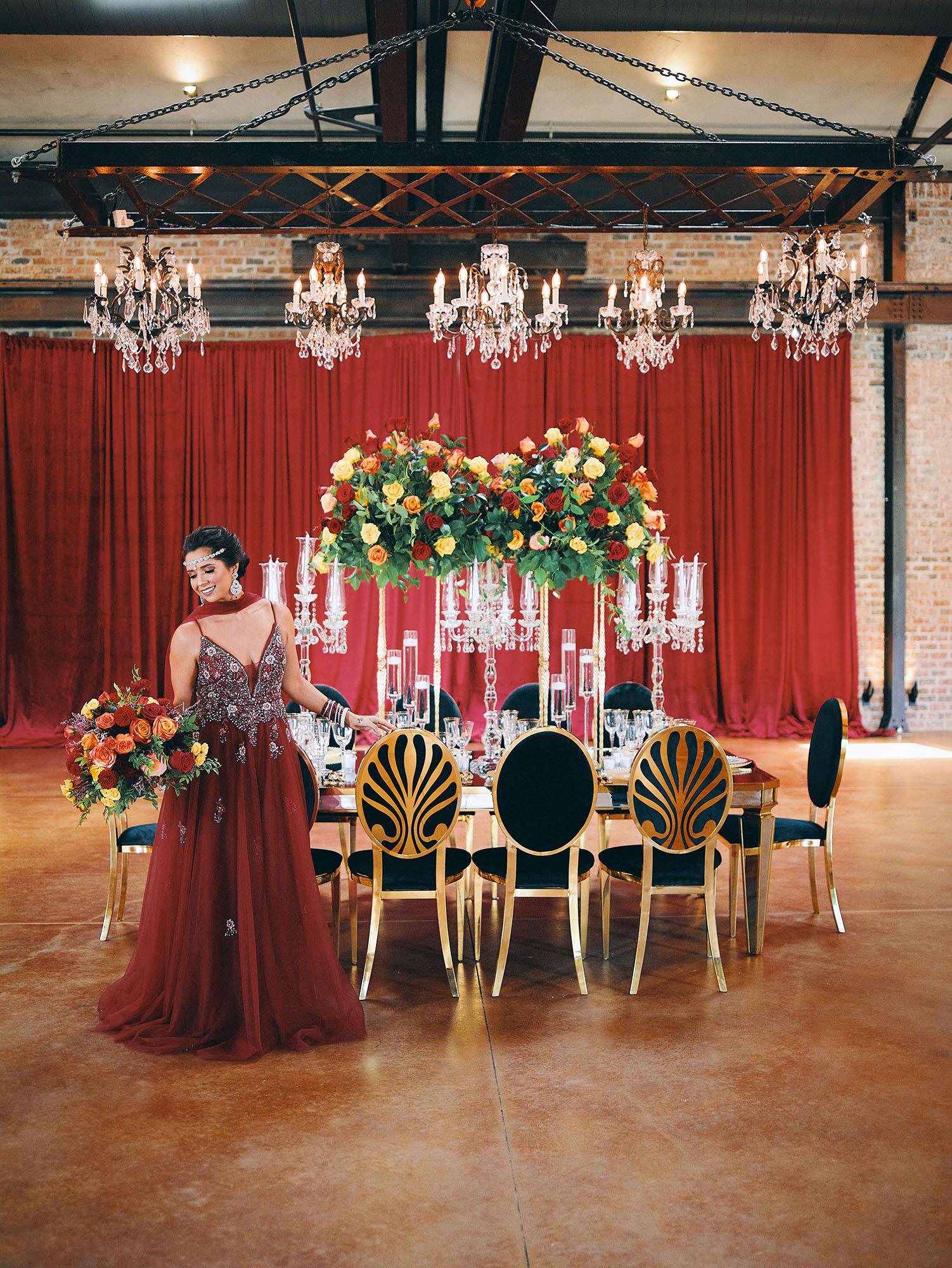 luxurious indoor wedding reception decor design, with red, yellow and orange floral designs, gold and black chairs, rectangular table, and a stunning bride and bridal bouquet.