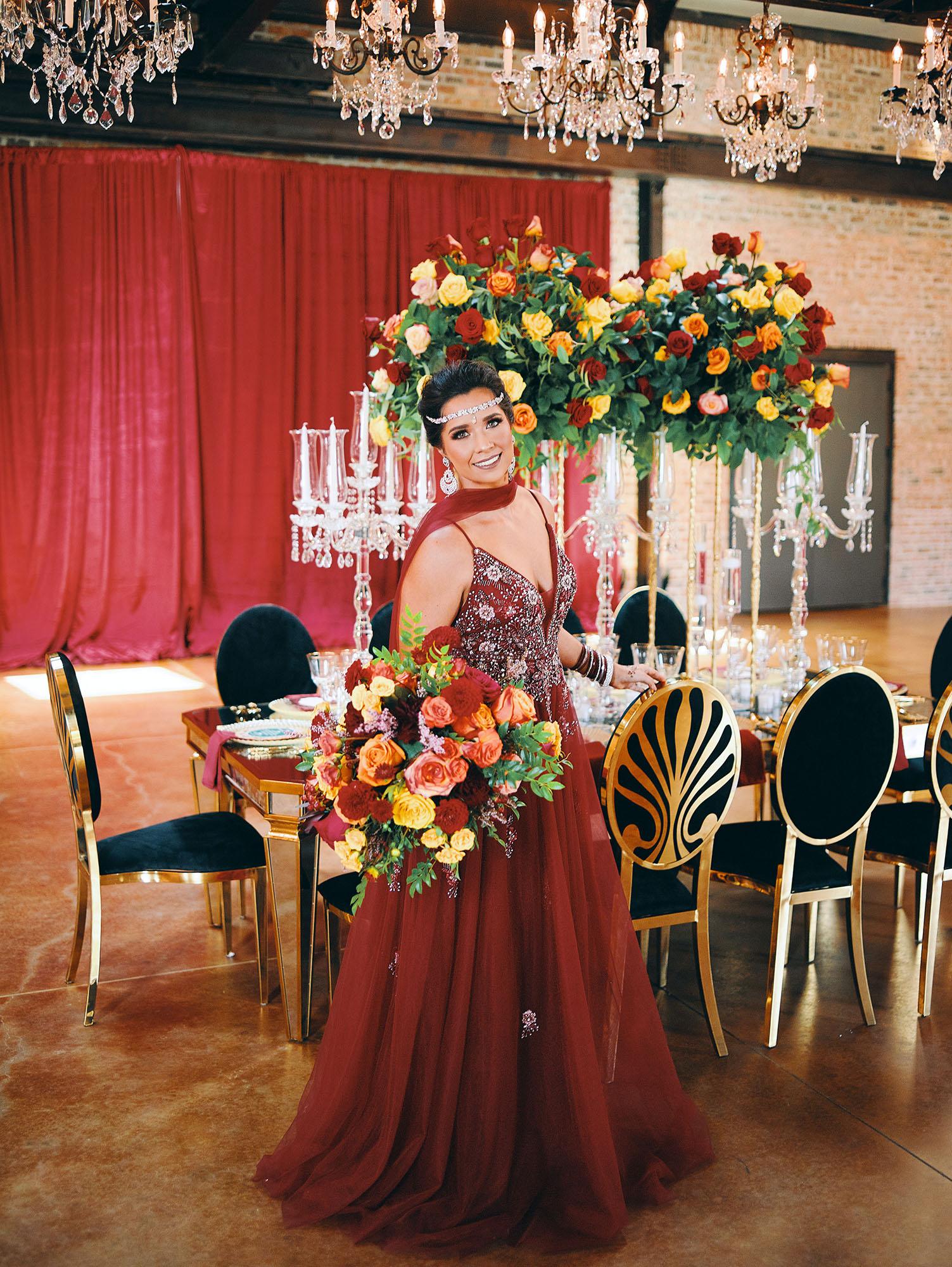 luxurious indoor wedding reception decor design, with red, yellow and orange floral designs, gold and black chairs, rectangular table, and a stunning bride and bridal bouquet.