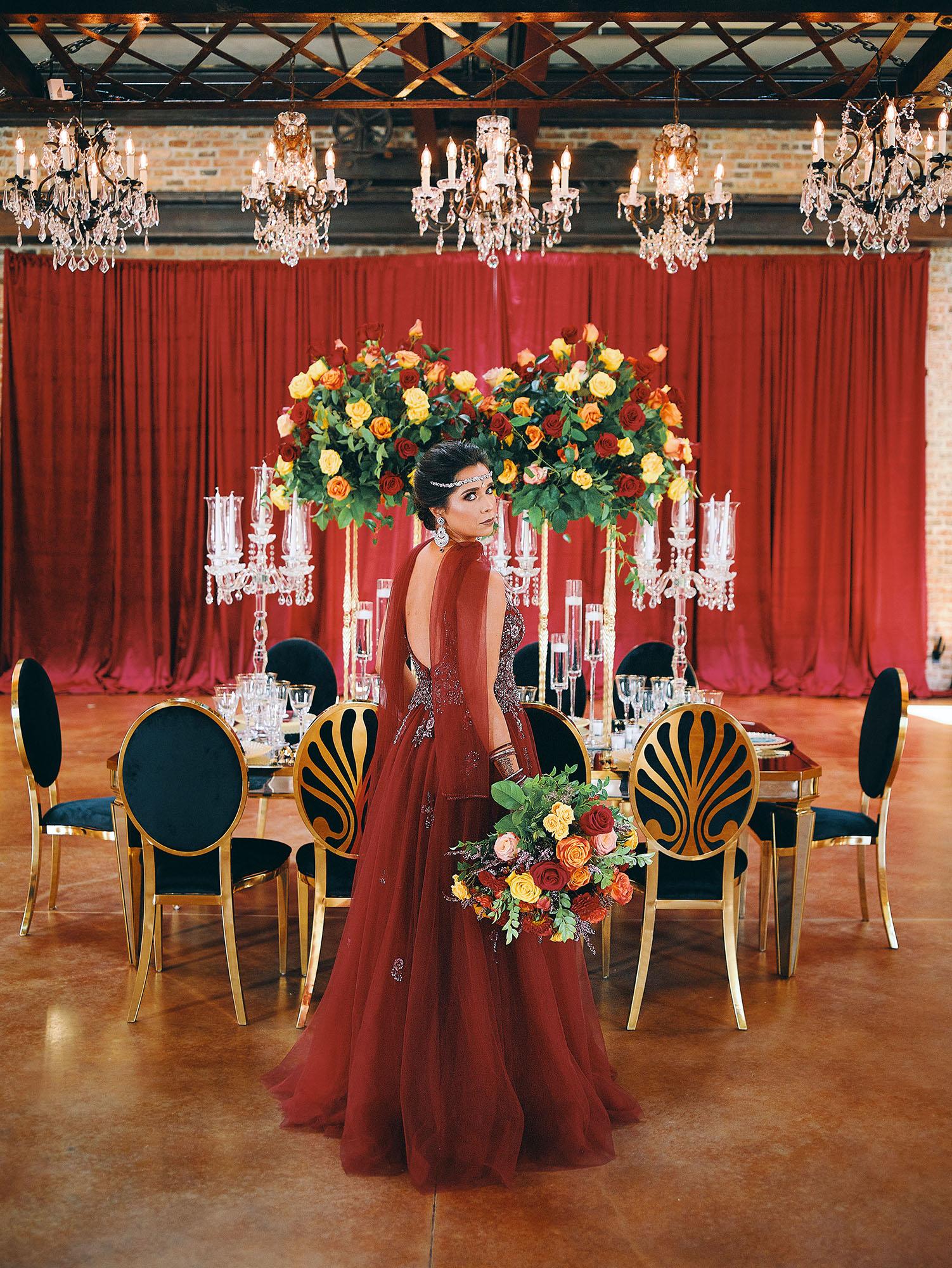 luxurious indoor wedding reception decor design, with red, yellow and orange floral designs, gold and black chairs, rectangular table, and a stunning bride and bridal bouquet.