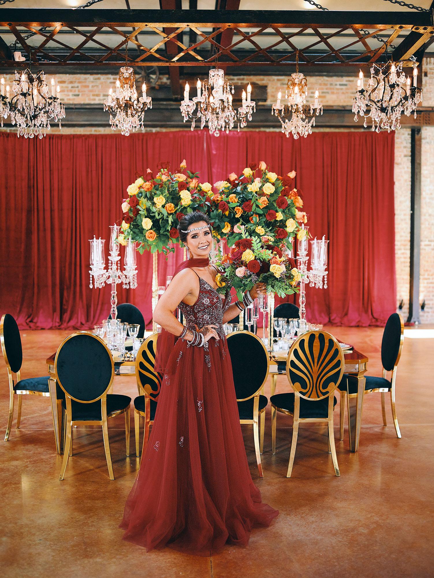 luxurious indoor wedding reception decor design, with red, yellow and orange floral designs, gold and black chairs, rectangular table, and a stunning bride and bridal bouquet.