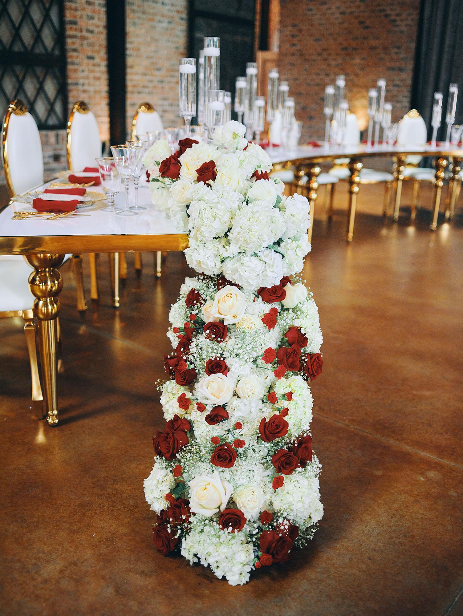 luxurious indoor wedding reception decor design, with red, and white floral designs, gold and white curved tables, gold chairs, and a stunning bride and bridal bouquet.
