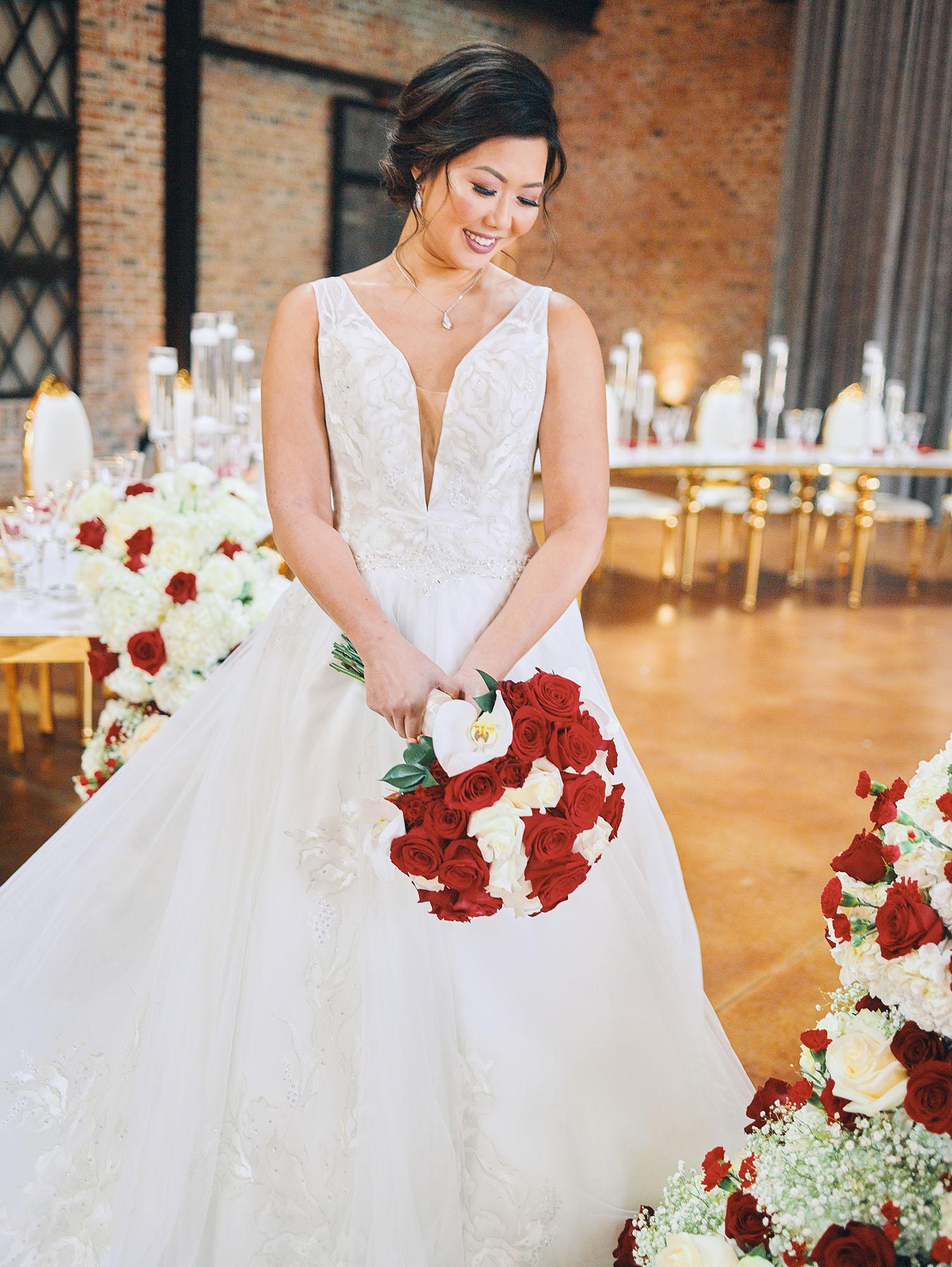 luxurious indoor wedding reception decor design, with red, and white floral designs, gold and white curved tables, gold chairs, and a stunning bride and bridal bouquet.