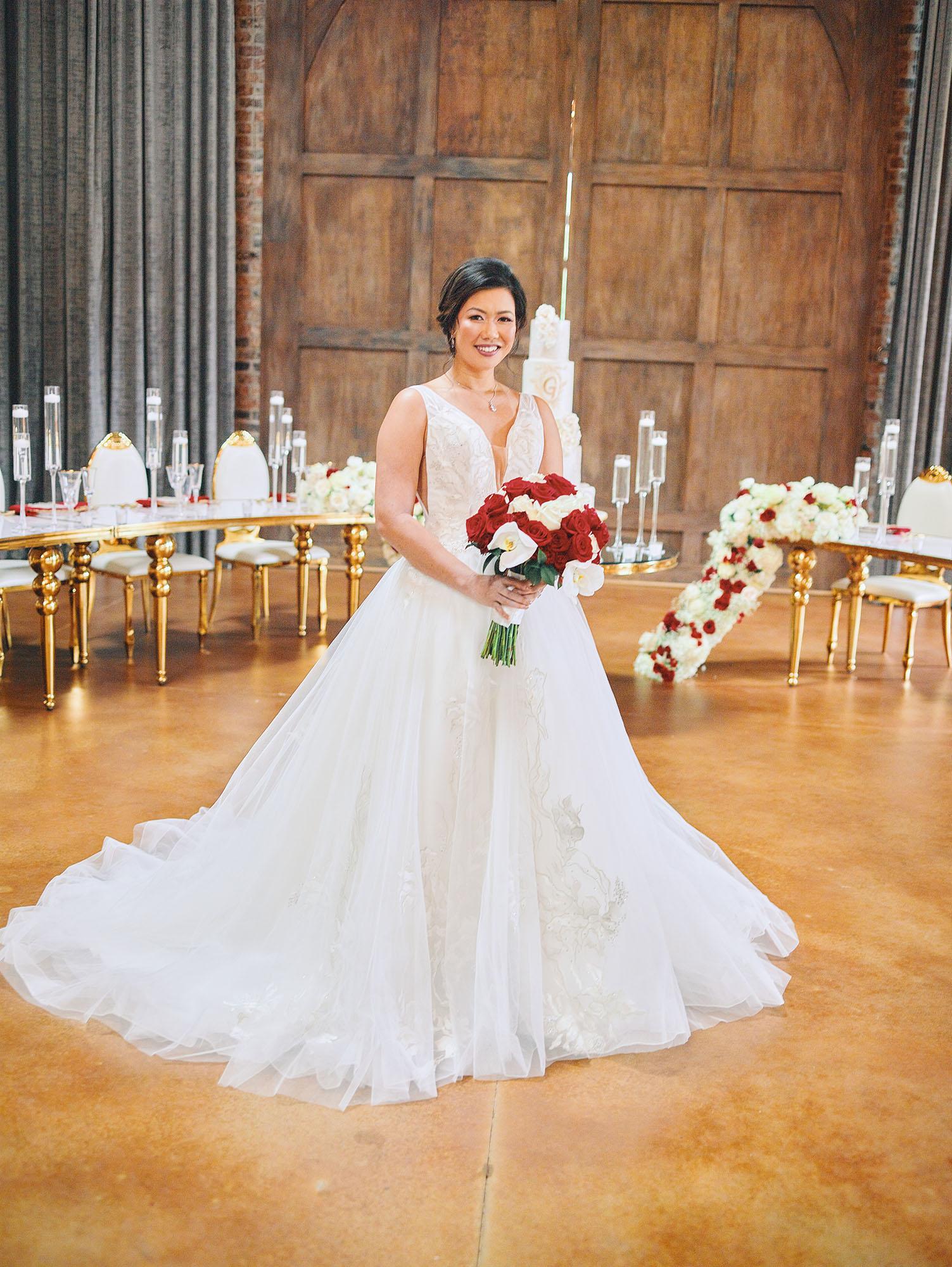 luxurious indoor wedding reception decor design, with red, and white floral designs, gold and white curved tables, gold chairs, and a stunning bride and bridal bouquet.
