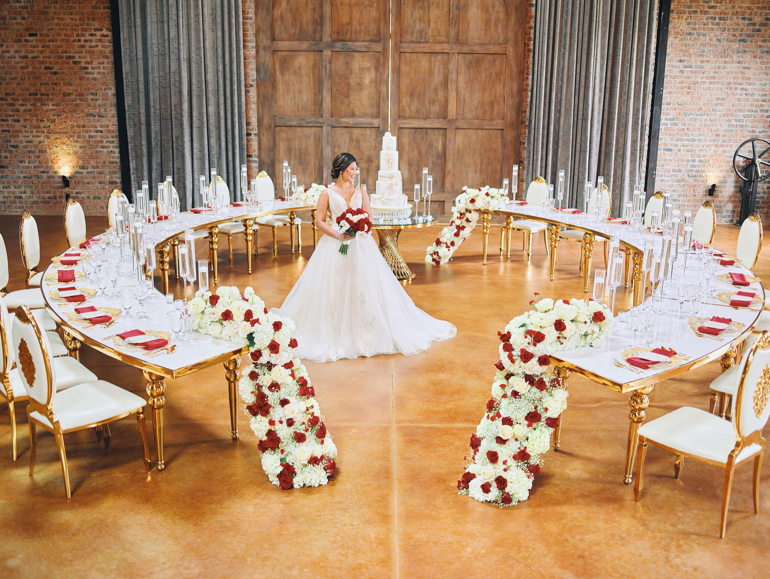 luxurious indoor wedding reception decor design, with red, and white floral designs, gold and white curved tables, gold chairs, and a stunning bride and bridal bouquet.