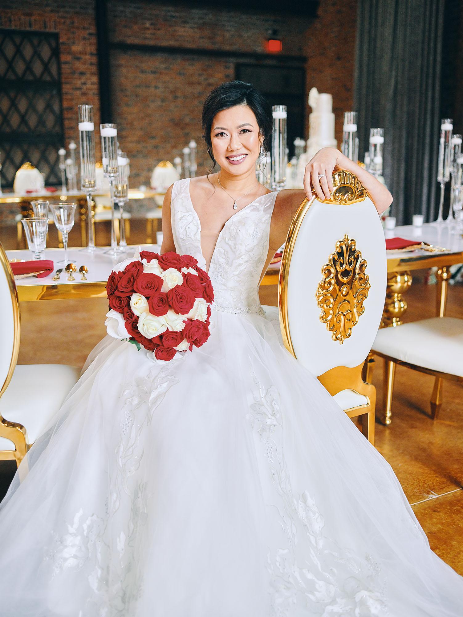 luxurious indoor wedding reception decor design, with red, and white floral designs, gold and white curved tables, gold chairs, and a stunning bride and bridal bouquet.