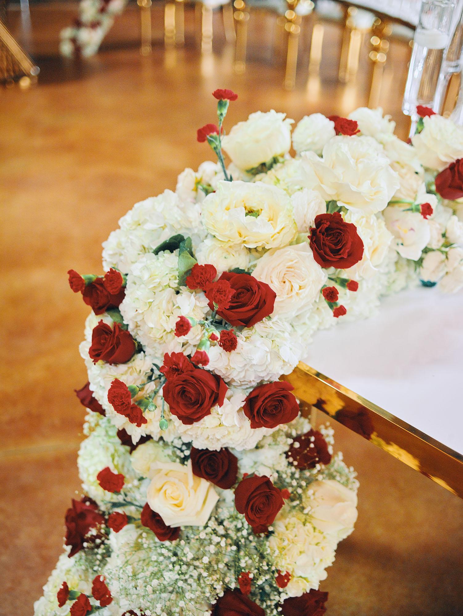 luxurious indoor wedding reception decor design, with red, and white floral designs, gold and white curved tables, gold chairs, and a stunning bride and bridal bouquet.