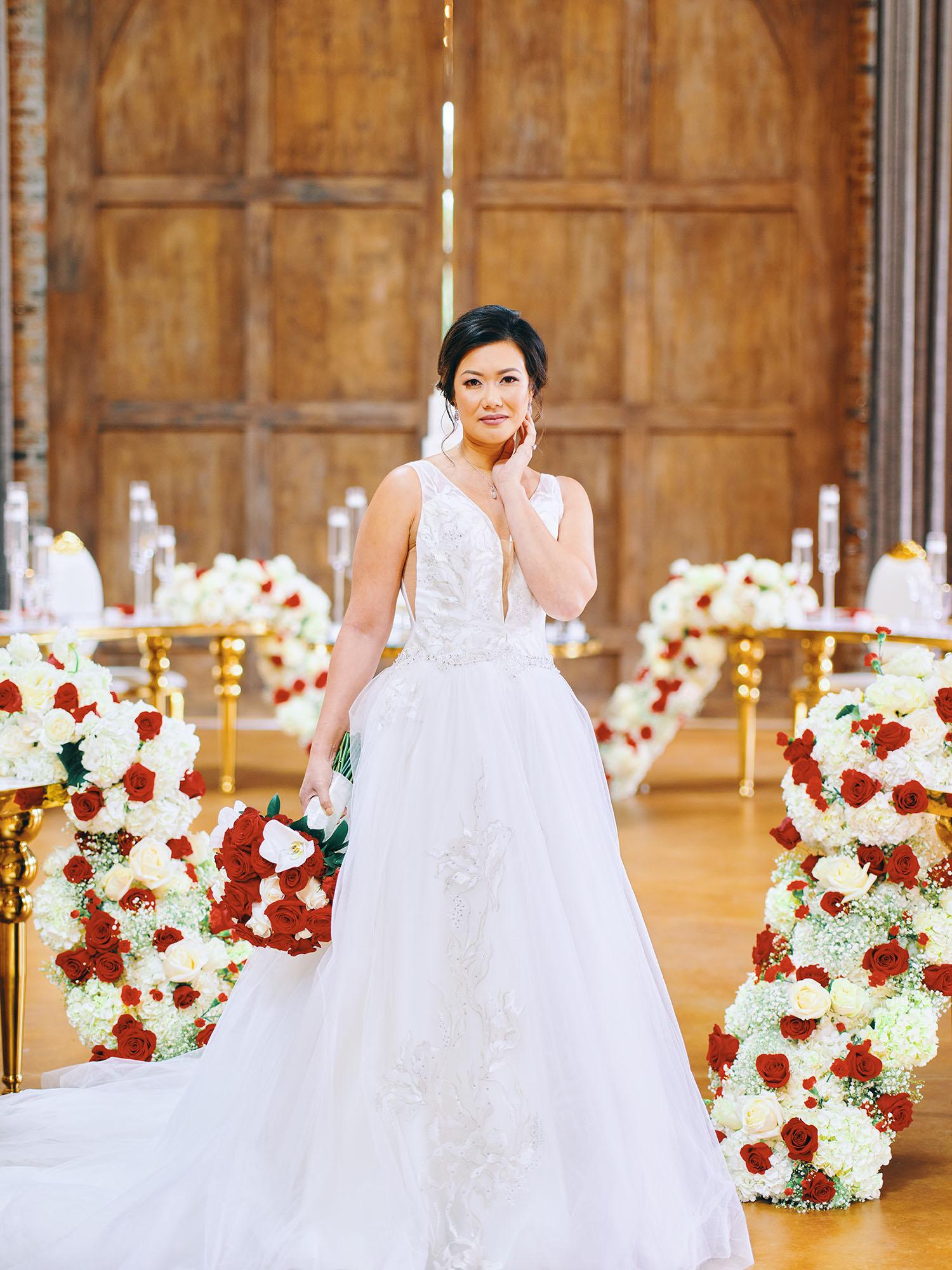 luxurious indoor wedding reception decor design, with red, and white floral designs, gold and white curved tables, gold chairs, and a stunning bride and bridal bouquet.