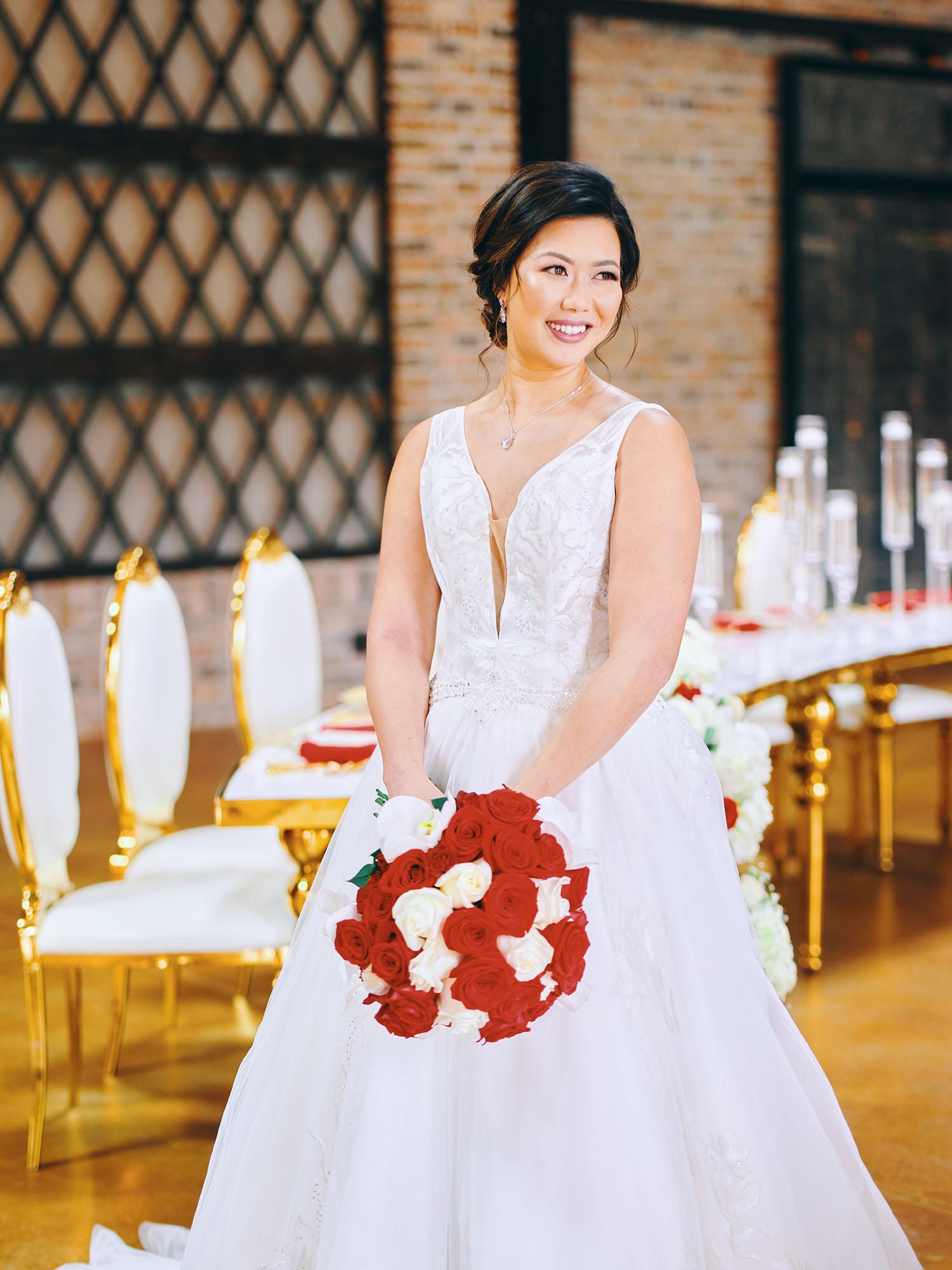 luxurious indoor wedding reception decor design, with red, and white floral designs, gold and white curved tables, gold chairs, and a stunning bride and bridal bouquet.