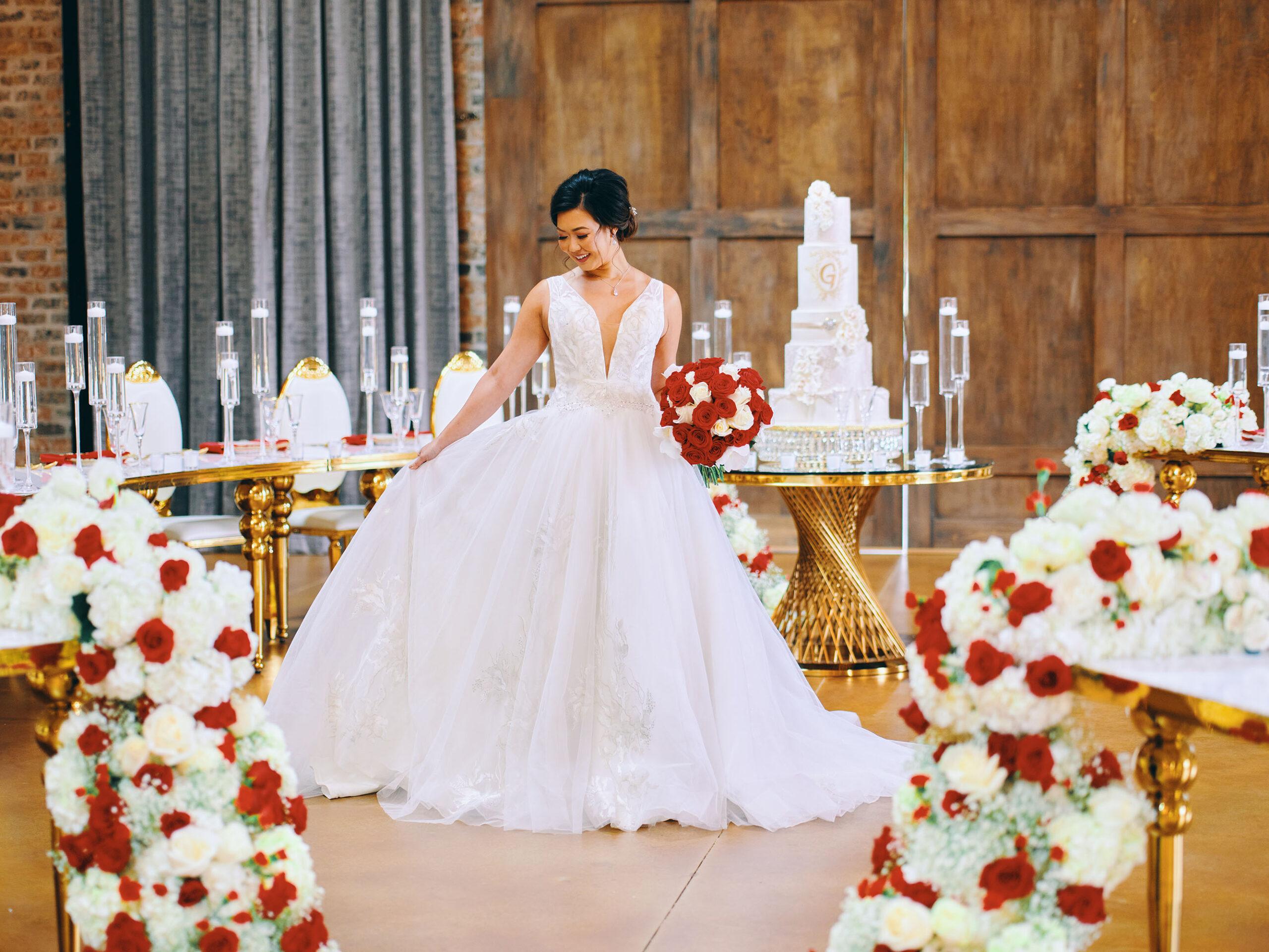 luxurious indoor wedding reception decor design, with red, and white floral designs, gold and white curved tables, gold chairs, and a stunning bride and bridal bouquet.