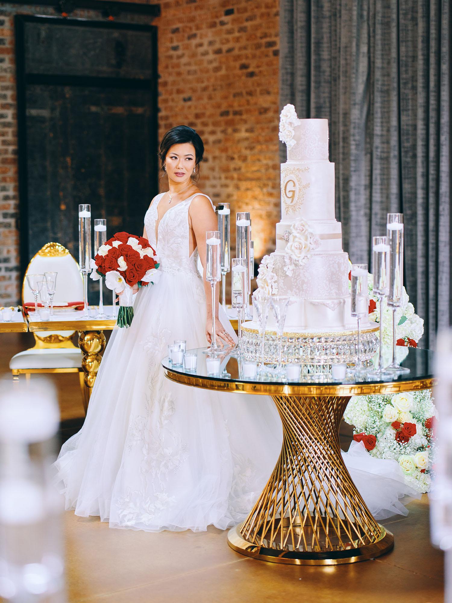 luxurious indoor wedding reception decor design, with red, and white floral designs, gold and white curved tables, gold chairs, and a stunning bride and bridal bouquet.