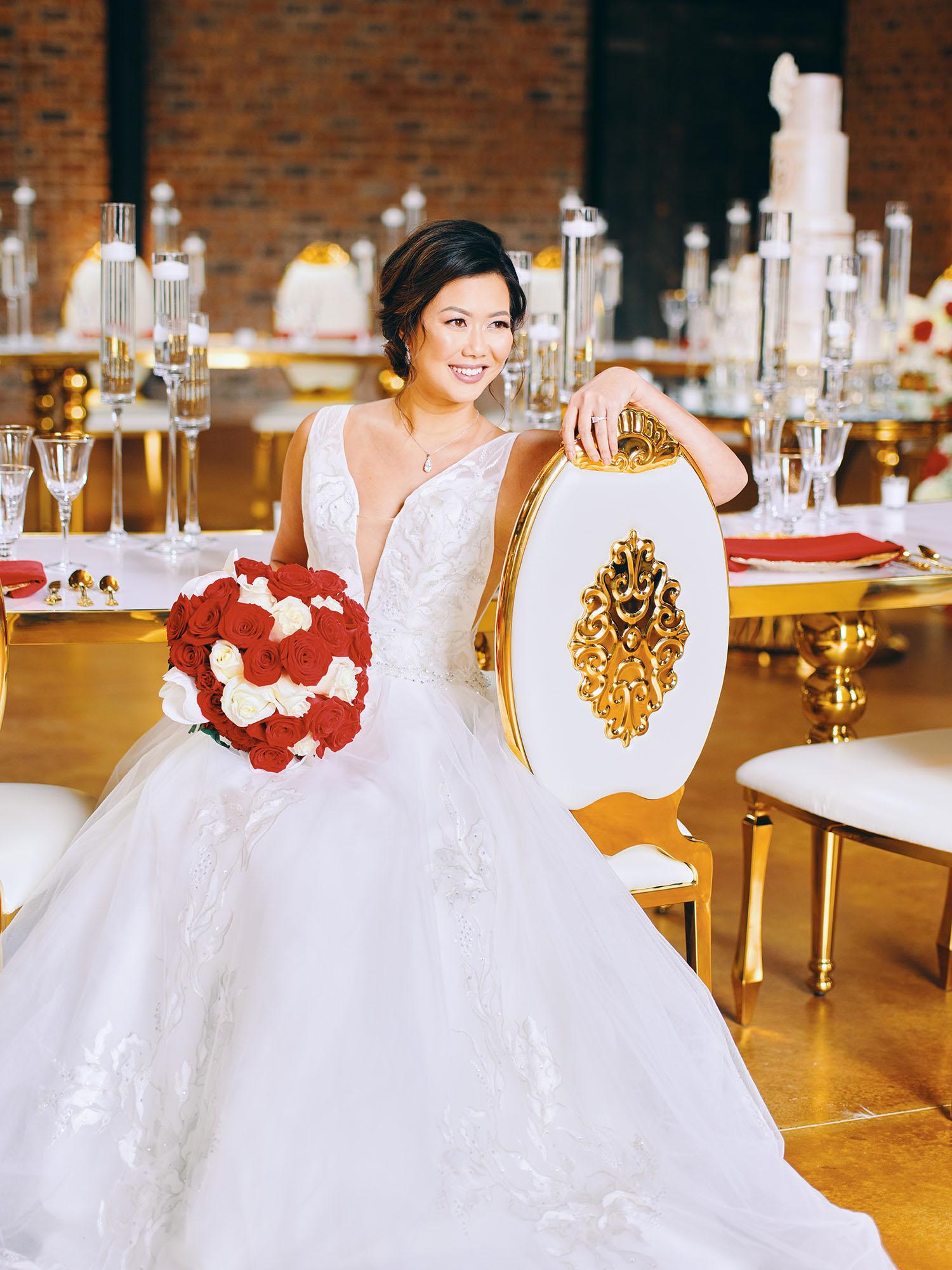 luxurious indoor wedding reception decor design, with red, and white floral designs, gold and white curved tables, gold chairs, and a stunning bride and bridal bouquet.